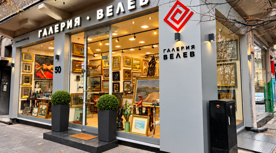 Art gallery storefront with a large window display of framed paintings and sculptures, sign in Cyrillic letters ГОГЛЕРИЯ ВЕЛЕВ above the entrance